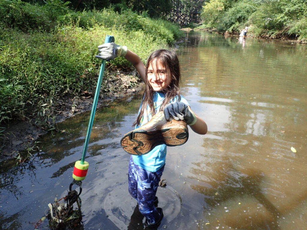 A young volunteer wading in a creek at a trash cleanup, holding an old shoe recovered from the water.